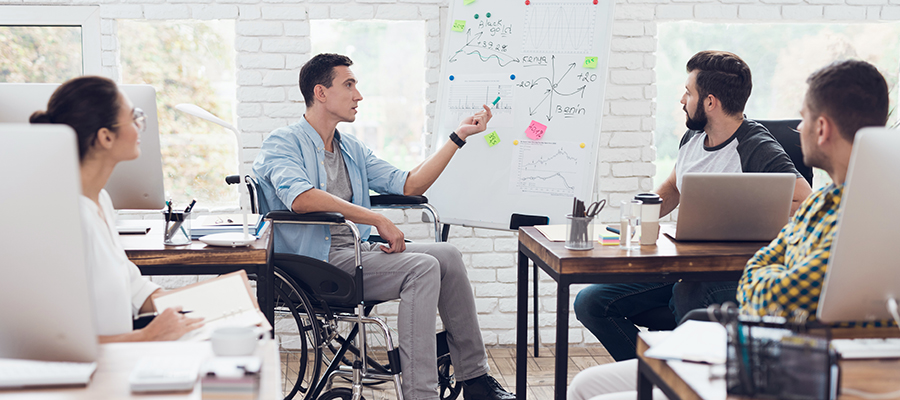 A man in a wheelchair demonstrates business graphics. He is holding a green marker. Collegues are listening attentively to him.