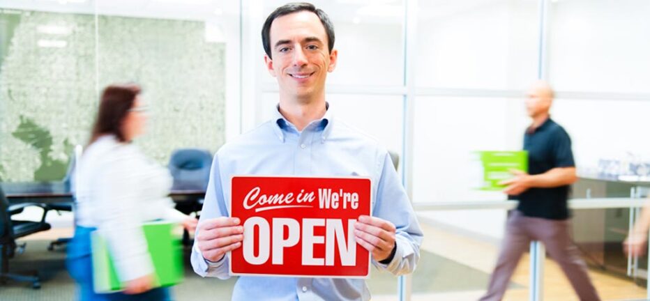Person standing in office setting holding sign that says Come in, we're open.